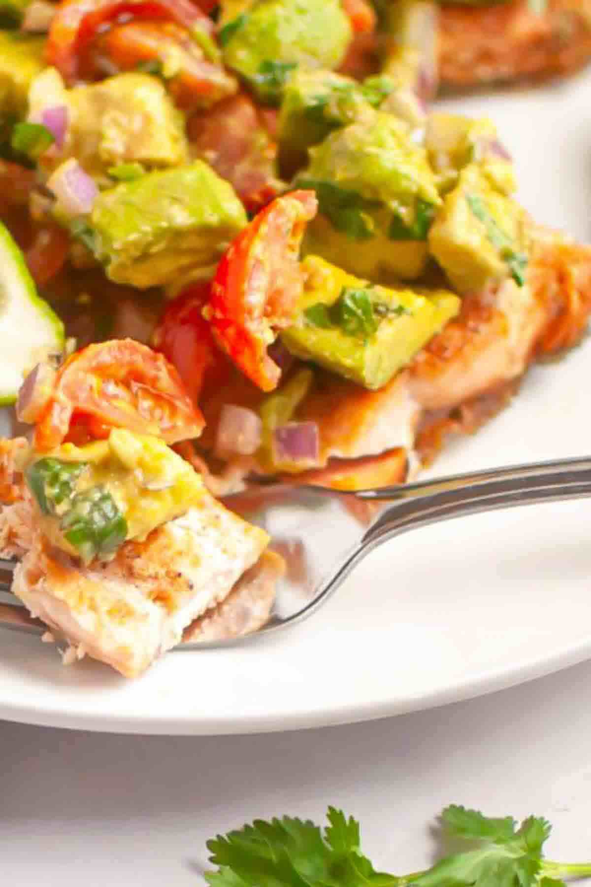 A fork holds a piece of grilled salmon topped with avocado, tomato, red onion, and cilantro salsa on a white plate.