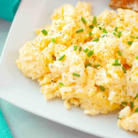 Scrambled eggs garnished with chopped chives and black pepper on a white plate, with a piece of toast visible in the corner.