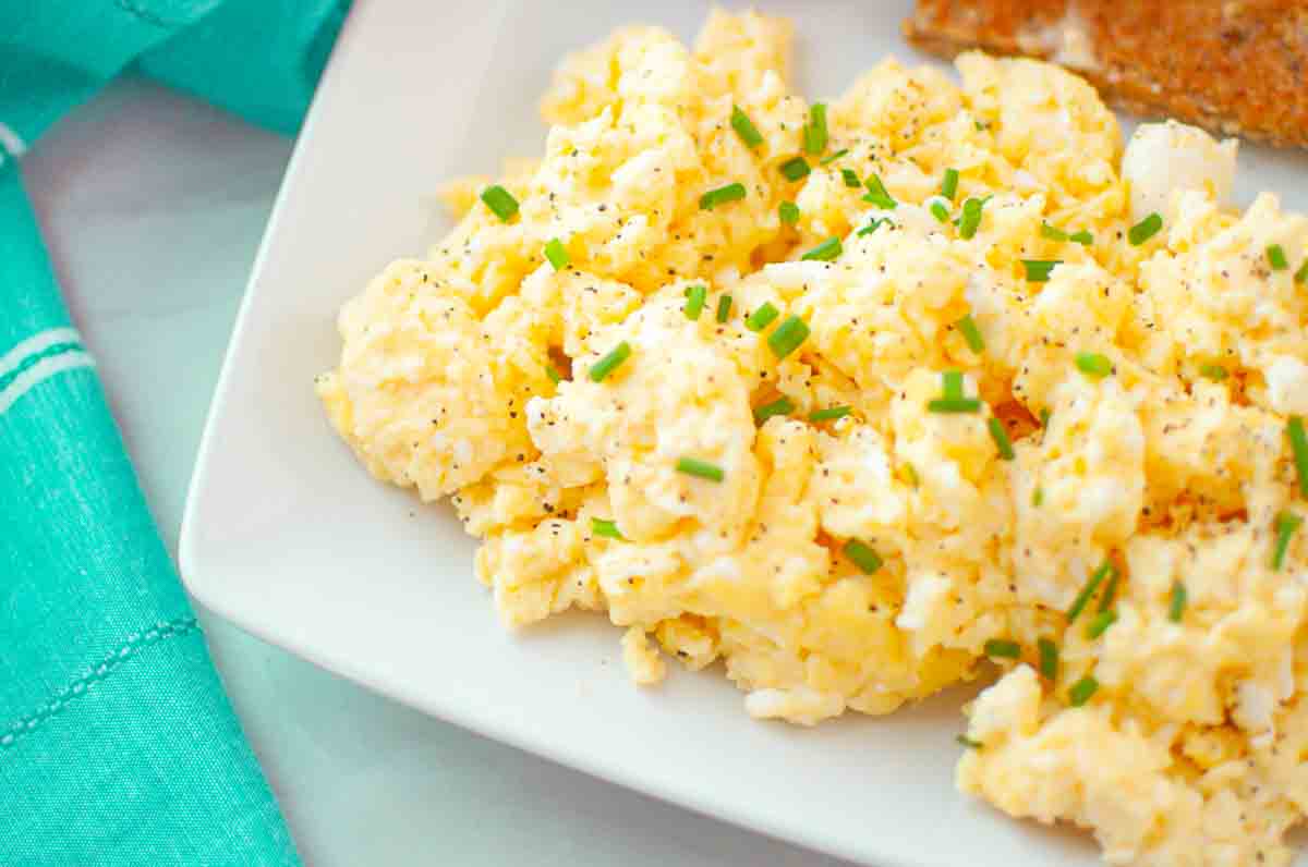 Scrambled eggs garnished with chopped chives and black pepper on a white plate, with a piece of toast visible in the corner.