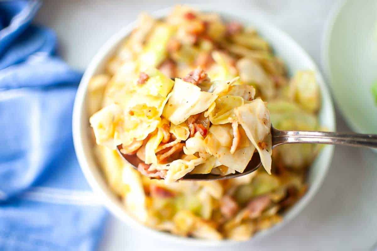 A spoonful of cooked cabbage mixed with pieces of bacon or ham held over a bowl filled with the same dish.