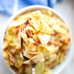 A close-up of a spoonful of cooked cabbage with pieces of bacon, held over a bowl filled with the same dish.