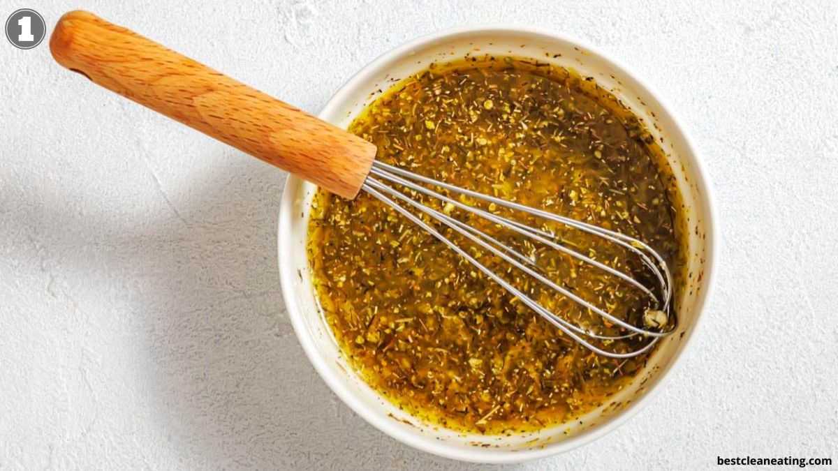A white bowl filled with a greenish-yellow herb dressing being mixed with a metal whisk with a wooden handle.