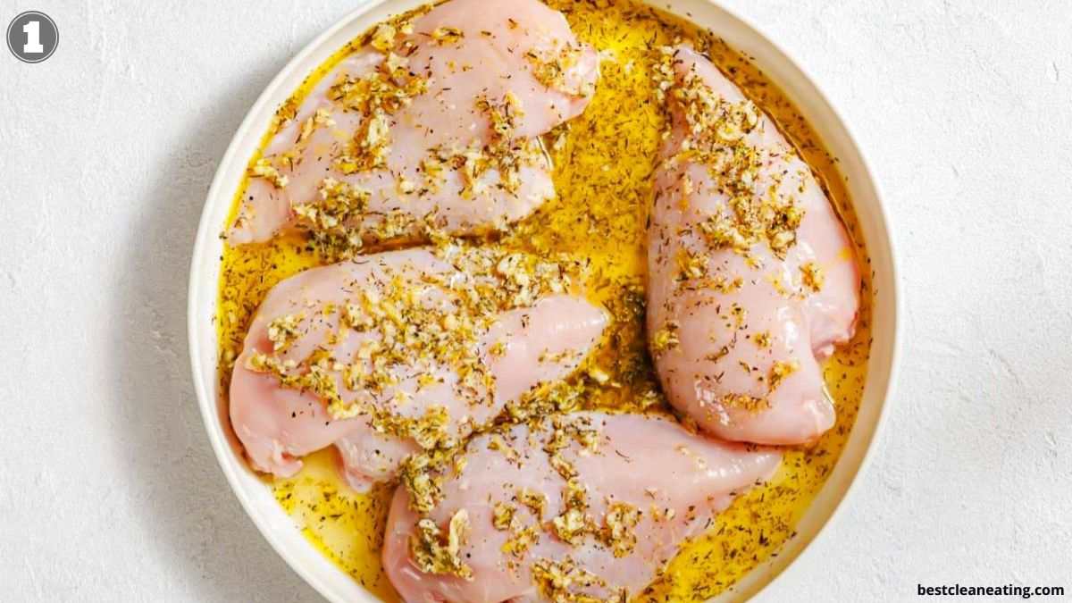 Raw chicken breasts marinating in a round dish with oil and herbs on a light background.