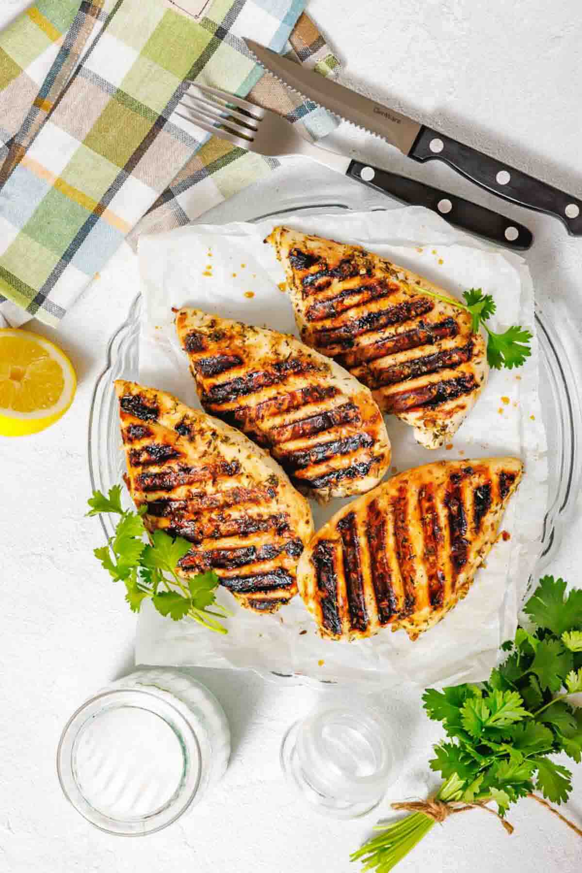 Four grilled chicken breasts with char marks on parchment paper, garnished with parsley, next to a lemon wedge, glass of water, and two knives on a checkered napkin.