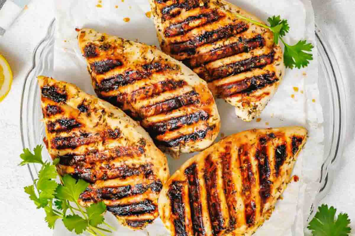 Four grilled chicken breasts with dark char marks are arranged on a white plate, garnished with fresh cilantro.