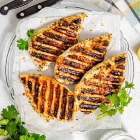 Four grilled chicken breasts with visible grill marks are arranged on parchment paper, garnished with fresh cilantro on a clear plate. Knives and a striped cloth are nearby.