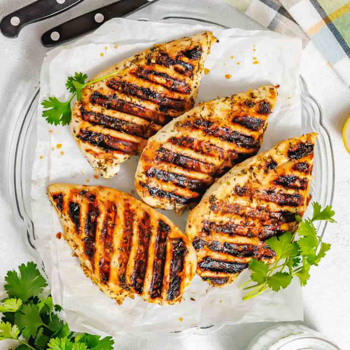 Four grilled chicken breasts with visible grill marks are arranged on parchment paper, garnished with fresh cilantro on a clear plate. Knives and a striped cloth are nearby.