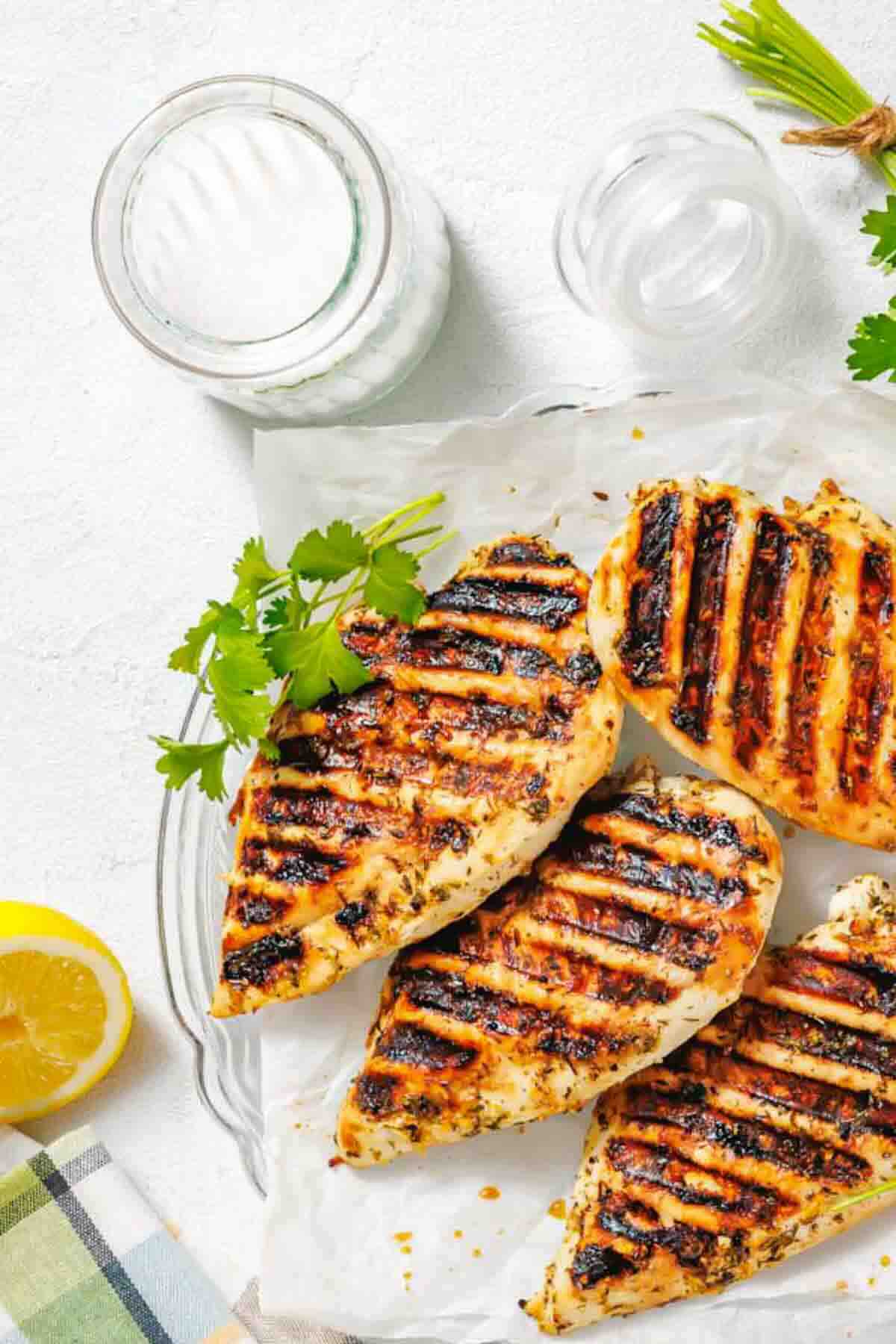 Three grilled chicken breasts with char marks on parchment paper, garnished with herbs, next to a glass of milk, a lemon half, and some fresh greens.
