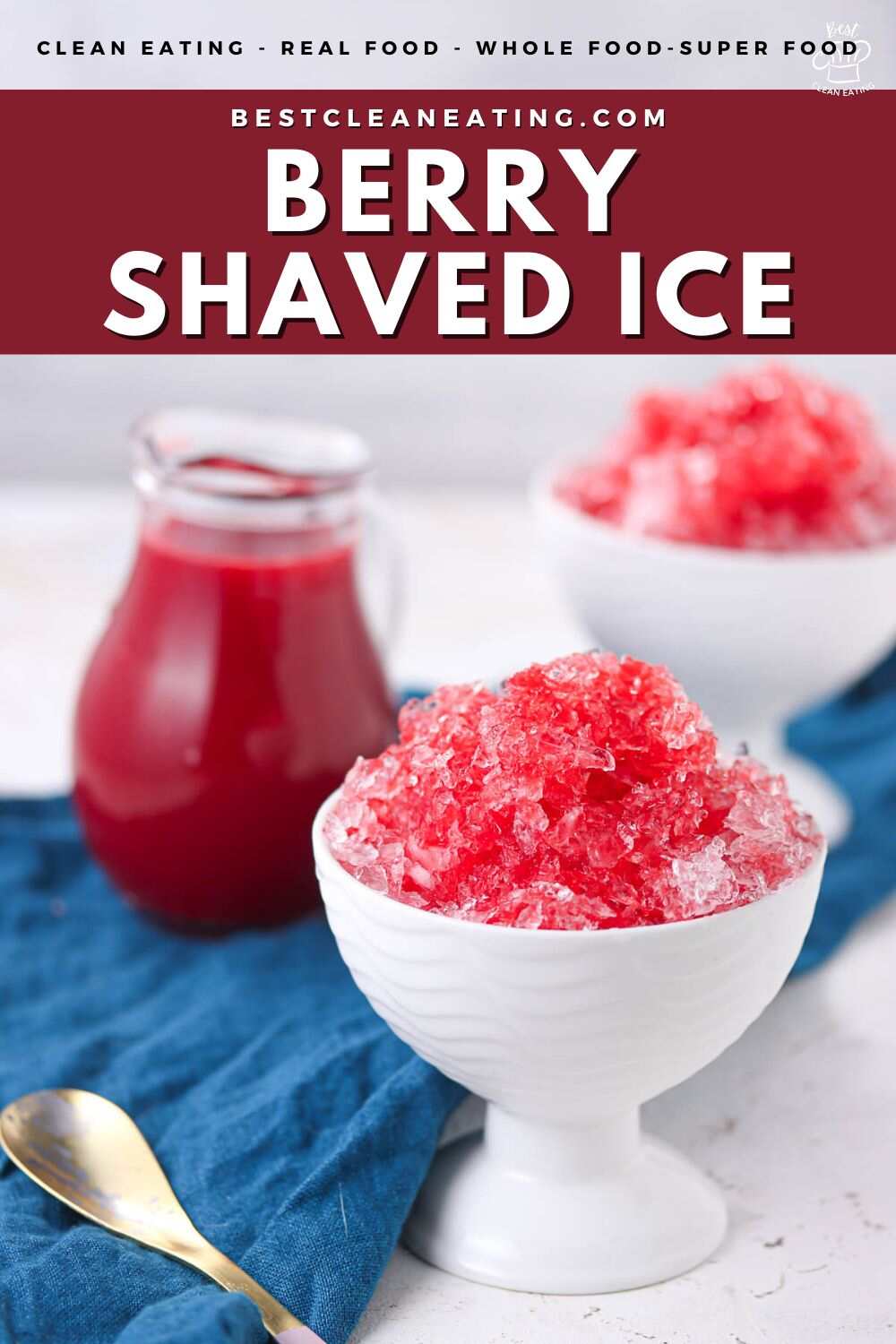 A white bowl filled with red berry shaved ice sits on a blue cloth next to a small glass pitcher of berry syrup and a gold spoon.