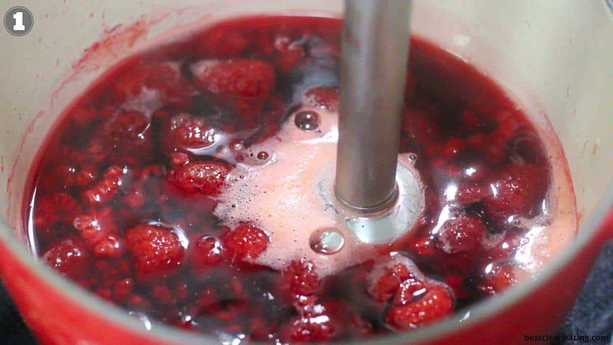 An immersion blender is blending red berries in a pot filled with liquid, creating a mixture with foam on the surface.
