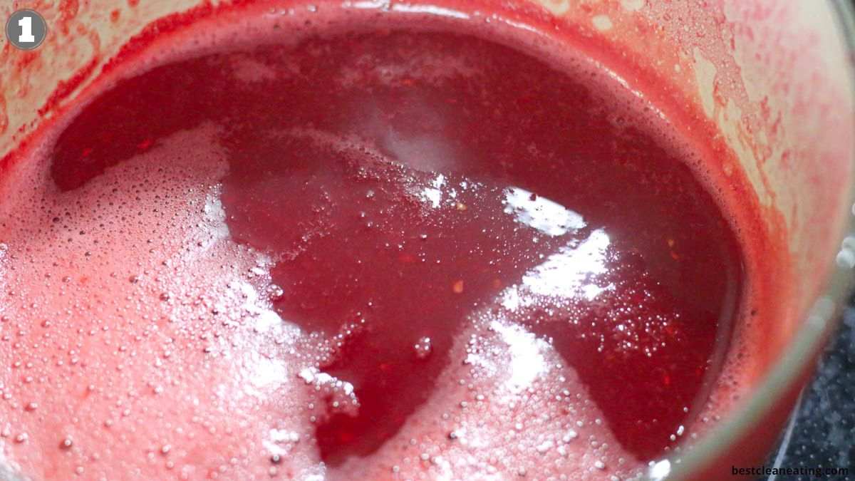A close-up view of a pot filled with simmering red liquid, likely a fruit sauce or syrup, with bubbles and foam on the surface.