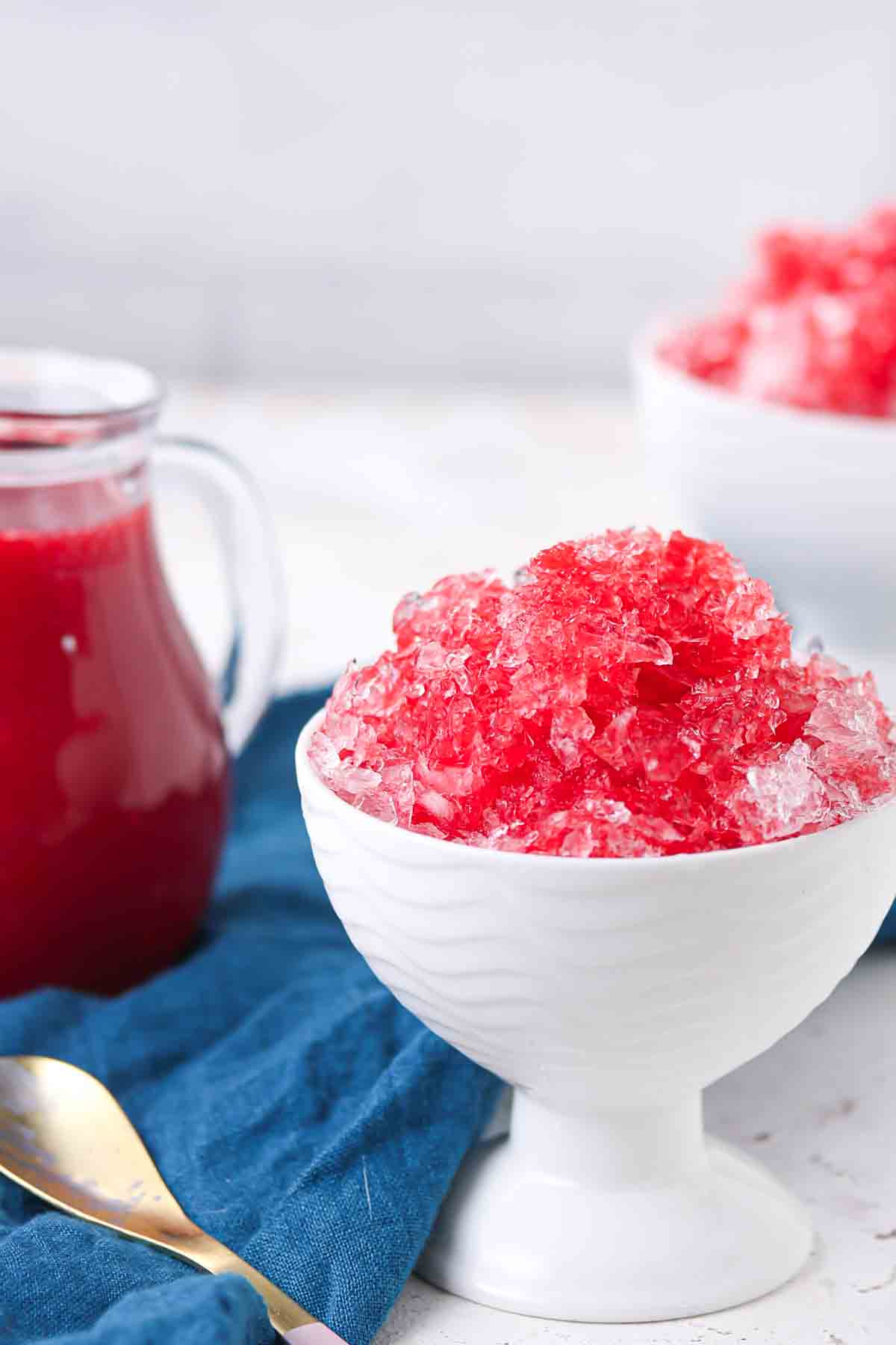 A white bowl filled with red shaved ice sits on a blue cloth, with a glass pitcher of red syrup and another bowl in the background.