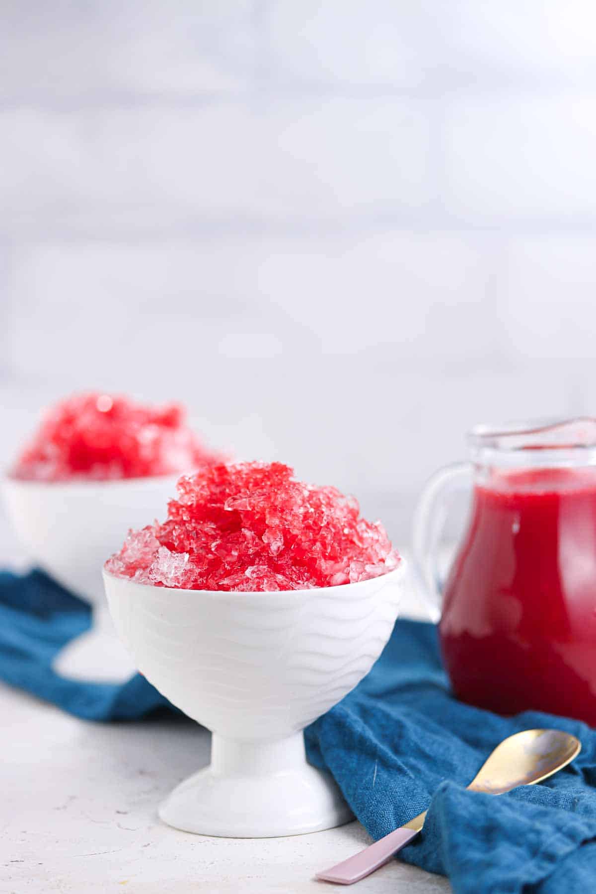 Two white bowls filled with red shaved ice sit on a blue cloth next to a glass pitcher of red syrup and a spoon.