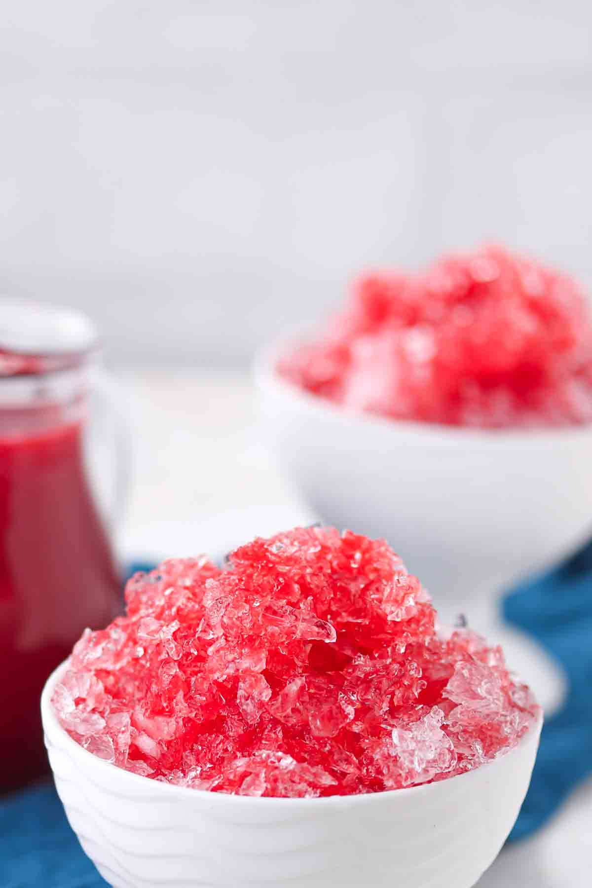 Two white bowls filled with red shaved ice sit on a blue napkin, with a glass container of red syrup in the background.