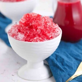 A white bowl filled with red shaved ice sits on a blue cloth, with a glass jar of red syrup in the background.