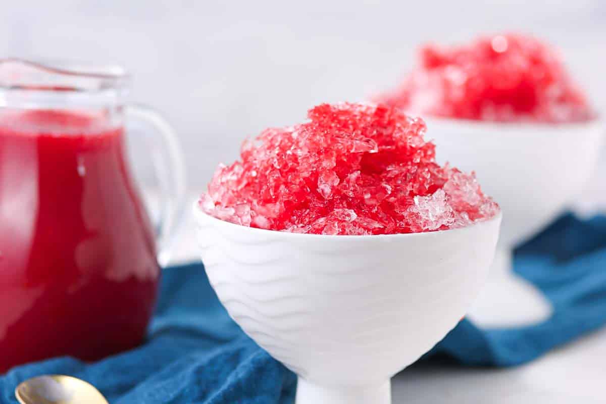 A white bowl filled with red shaved ice sits on a blue cloth, with a glass pitcher of red syrup and another bowl of shaved ice in the background.
