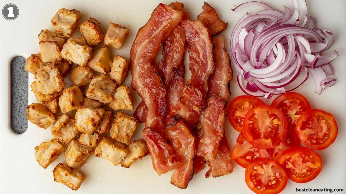 Chopped cooked chicken, strips of bacon, sliced red onions, and sliced tomatoes arranged on a white cutting board.