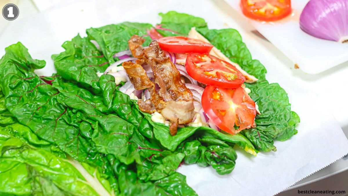A close-up of a leaf wrap with cooked meat, sliced tomatoes, and onions on green leafy greens, with sliced tomatoes and a red onion partially visible in the background.