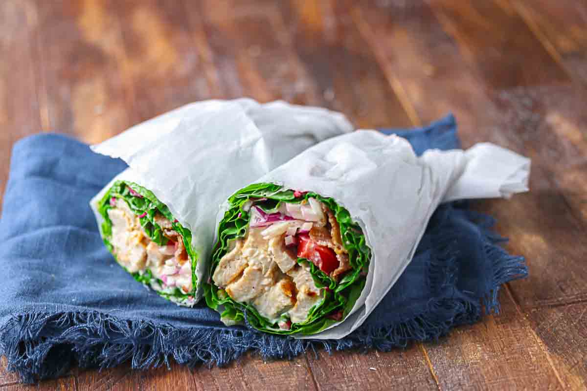 Two lettuce wraps filled with grilled chicken, sliced red onions, and tomatoes, wrapped in white parchment paper, placed on a blue cloth over a wooden surface.