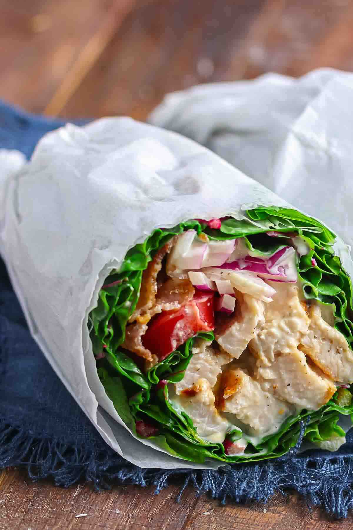 A close-up of a lettuce wrap filled with grilled chicken, tomatoes, onions, and sauce, partially wrapped in white parchment paper on a dark cloth.