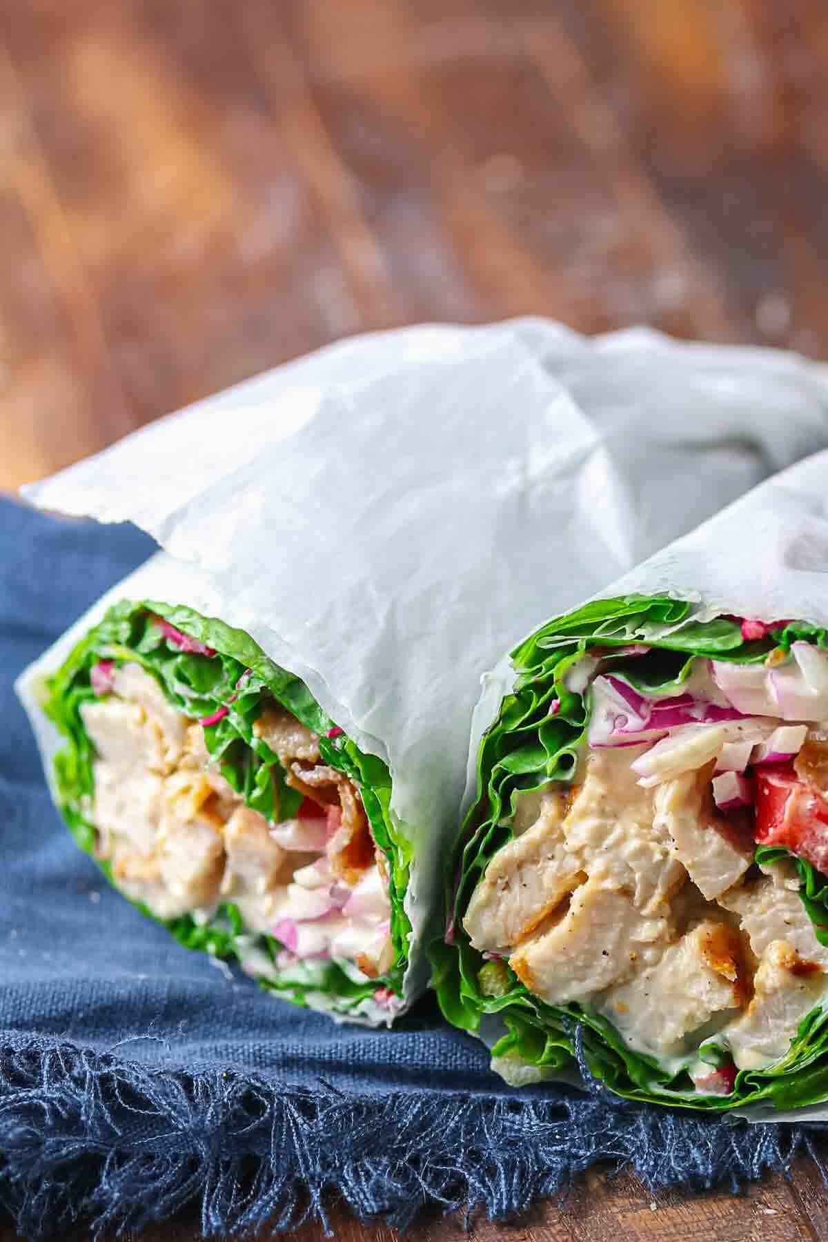 Two lettuce wraps filled with grilled chicken, tomatoes, red onion, and cabbage, wrapped in parchment paper and placed on a blue cloth.