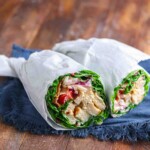 Two lettuce wraps filled with grilled chicken, tomatoes, onions, and other vegetables, cut in half and wrapped in white paper, placed on a blue cloth on a wooden surface.
