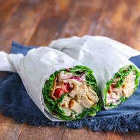 Two lettuce wraps filled with grilled chicken, tomatoes, onions, and other vegetables, cut in half and wrapped in white paper, placed on a blue cloth on a wooden surface.