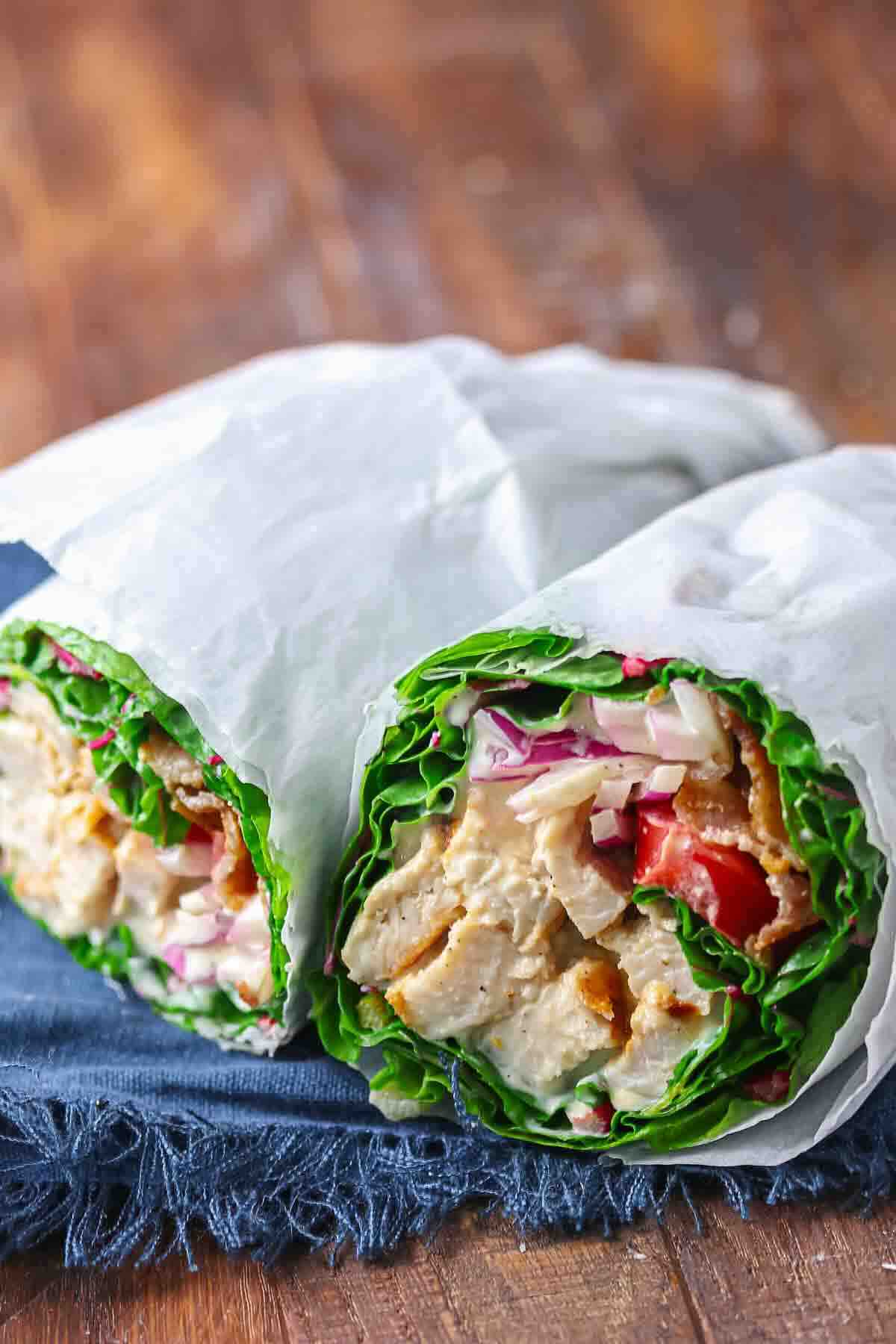 Two lettuce wraps filled with chicken, tomatoes, red onions, and cheese, partially wrapped in white paper and placed on a dark blue cloth.