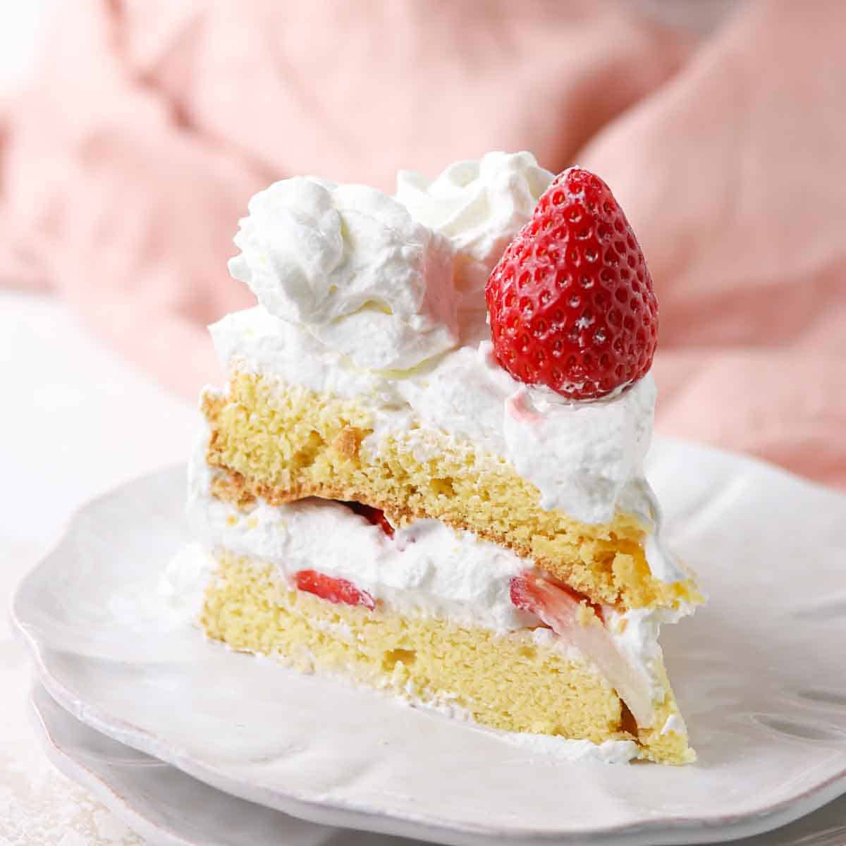 A slice of layered sponge cake with whipped cream and strawberries on top, served on a white plate.