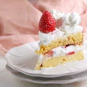 A slice of strawberry shortcake with whipped cream on a white plate, topped with a whole strawberry.