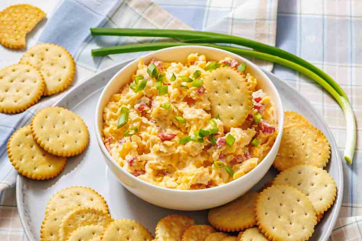 A bowl of pimento cheese dip garnished with chopped green onions, surrounded by round crackers on a plate.