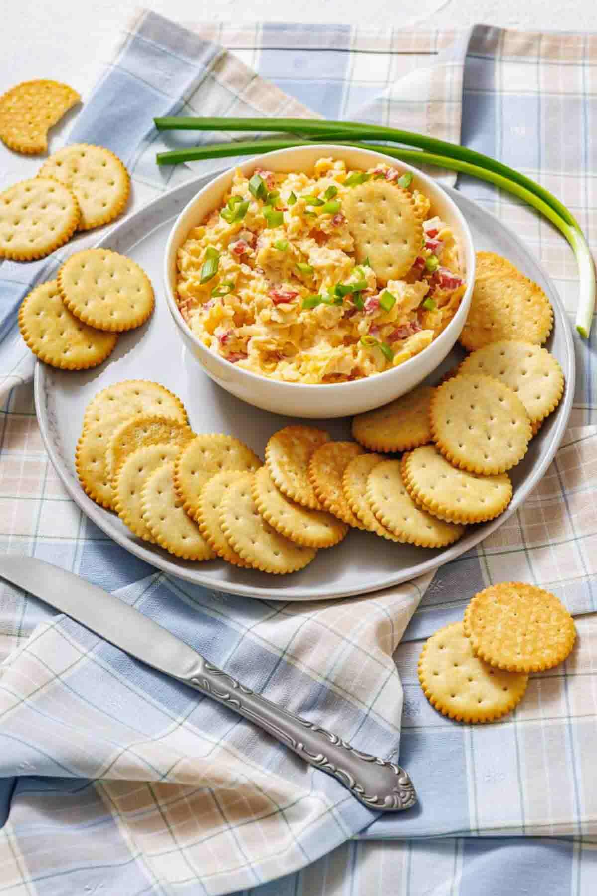 A bowl of pimento cheese dip topped with chopped green onions, surrounded by round crackers on a plate, with a knife and green onions on a plaid cloth.