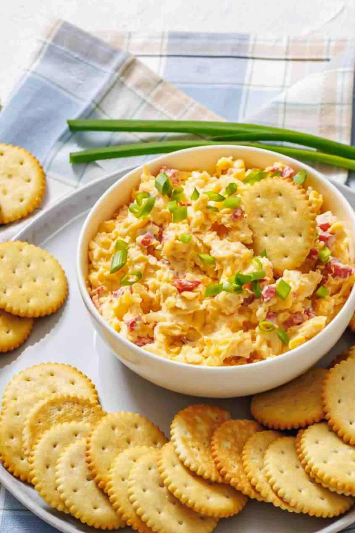 A bowl of pimento cheese dip topped with chopped green onions, surrounded by round crackers on a white plate.