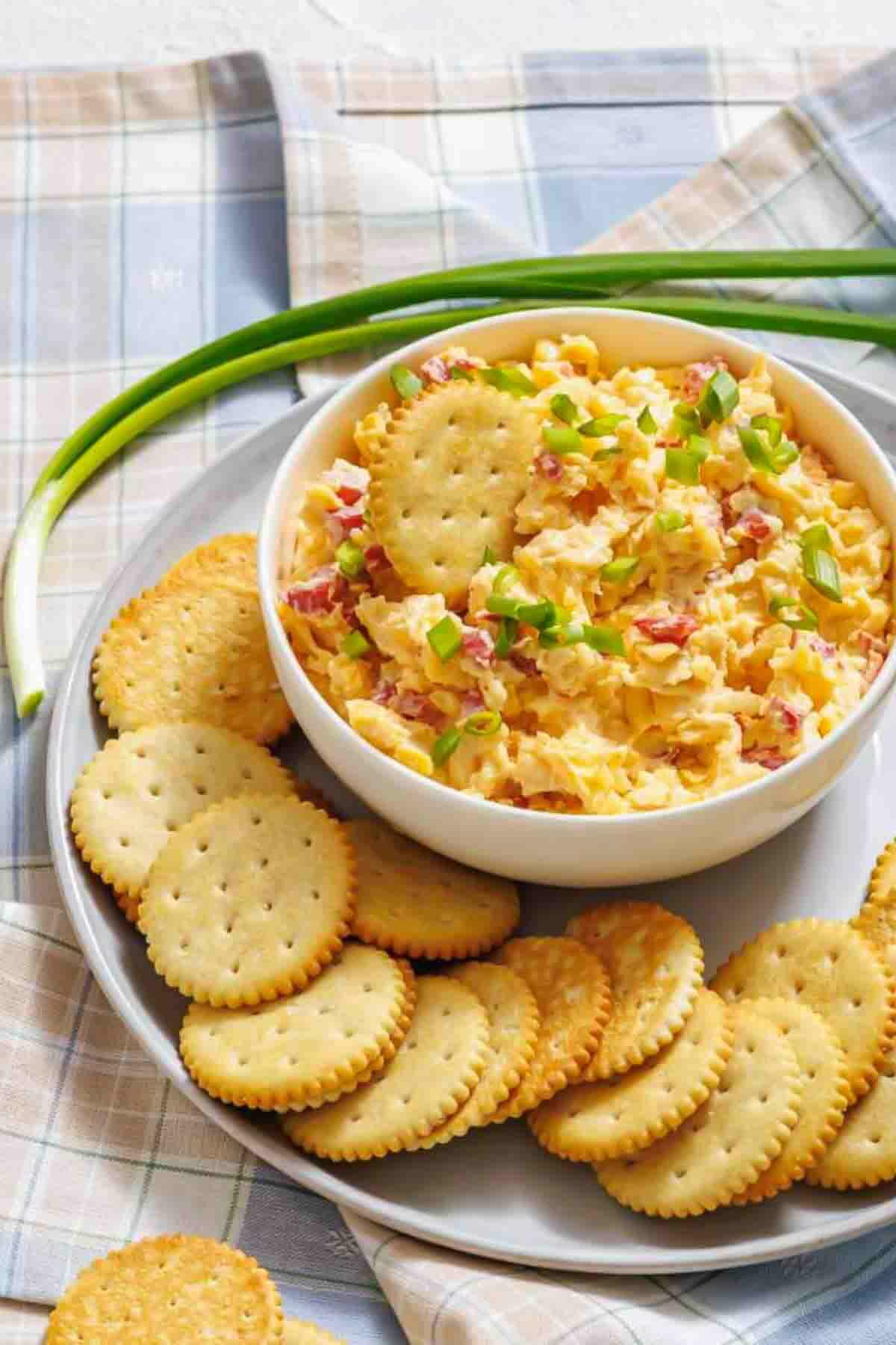 A bowl of pimento cheese dip topped with chopped green onions, surrounded by round crackers on a plate, with green onions in the background.