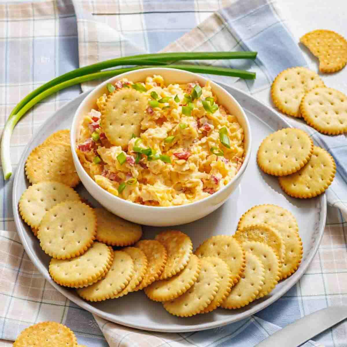 A bowl of pimento cheese dip garnished with chopped green onions is surrounded by round crackers on a plate, with whole green onions beside it.