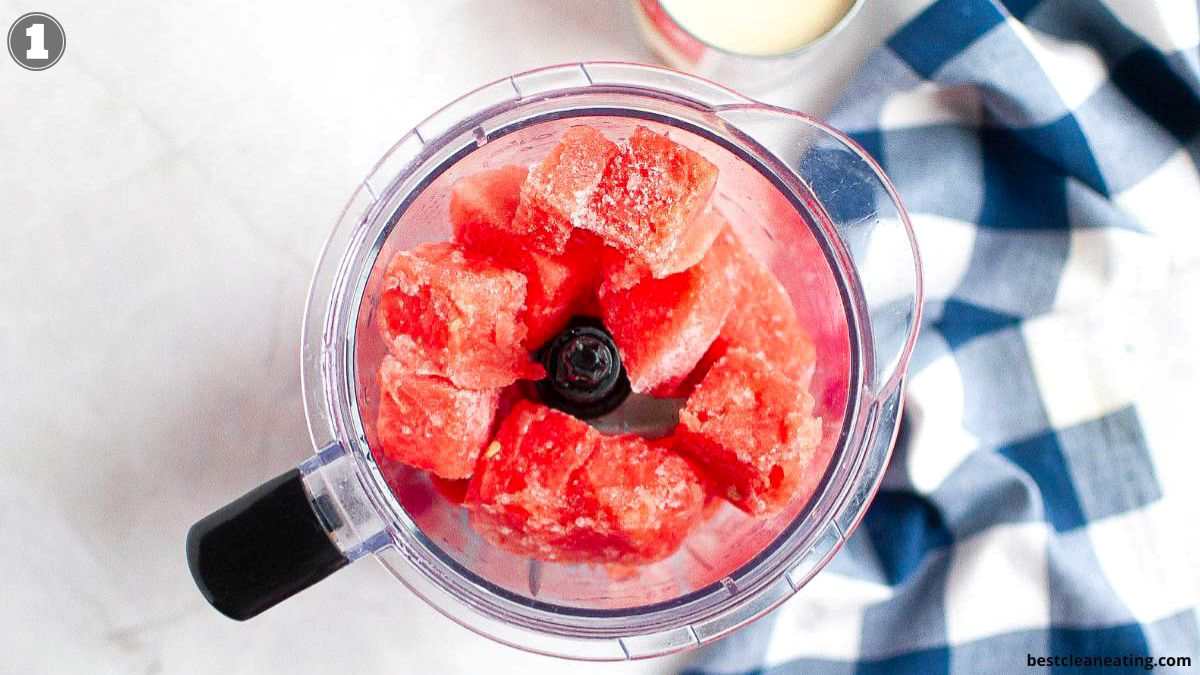 A top-down view of a blender filled with frozen watermelon chunks, placed on a white surface next to a blue and white checkered cloth.
