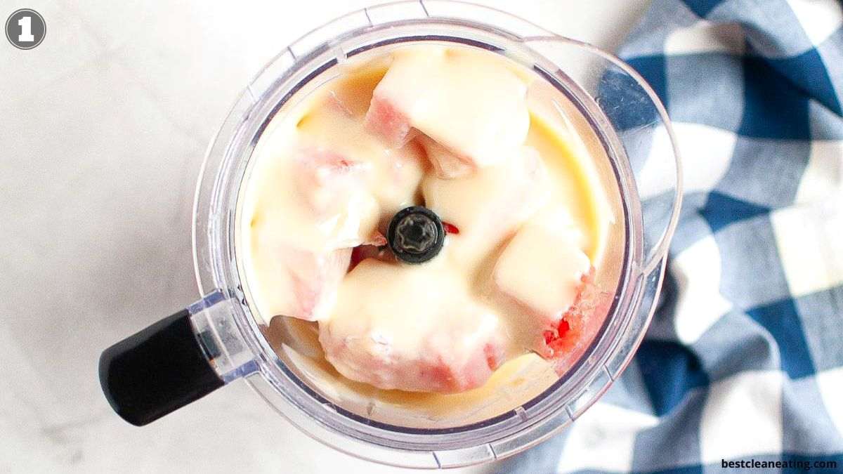 A blender filled with chunks of fruit and a creamy liquid, viewed from above, next to a blue and white checkered cloth.