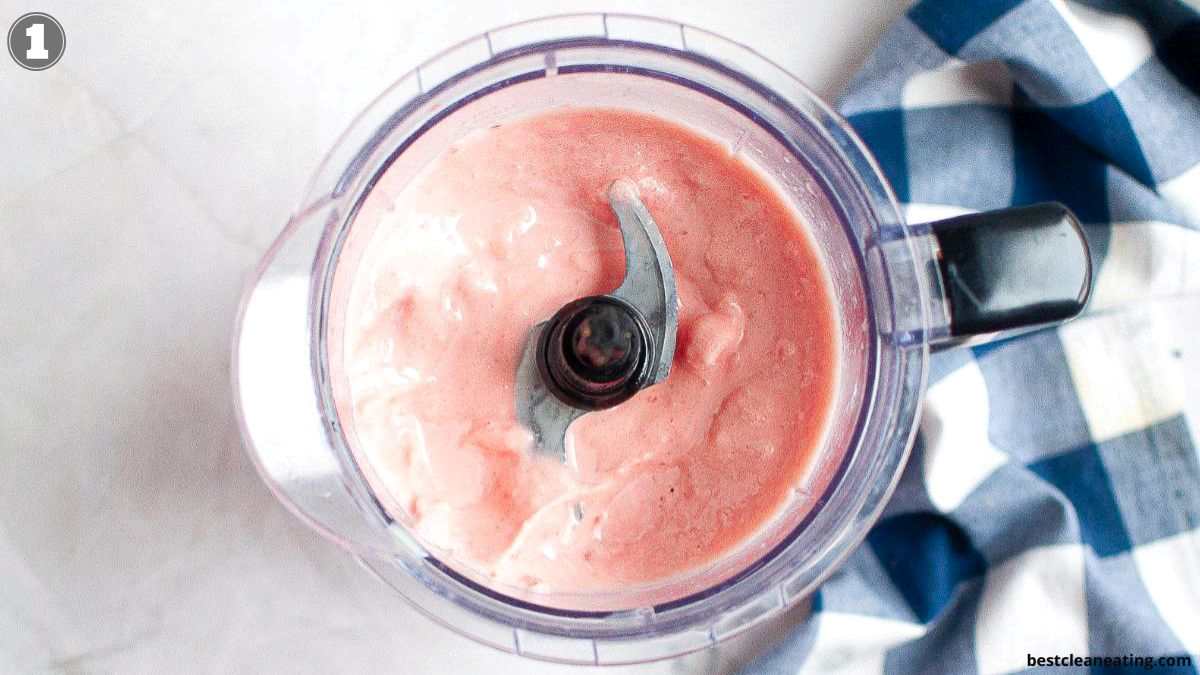 A blender with a pink creamy mixture inside, viewed from above, next to a blue and white checkered cloth.