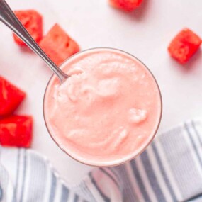 A glass of pink watermelon smoothie with a spoon, surrounded by watermelon cubes on a white surface with a striped cloth.