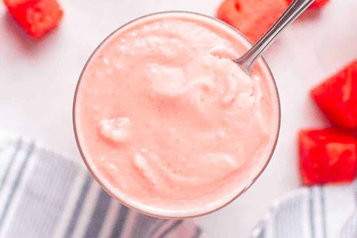 A glass filled with a smooth, pink frozen sherbet, with a spoon inside. Pieces of watermelon are visible in the background.
