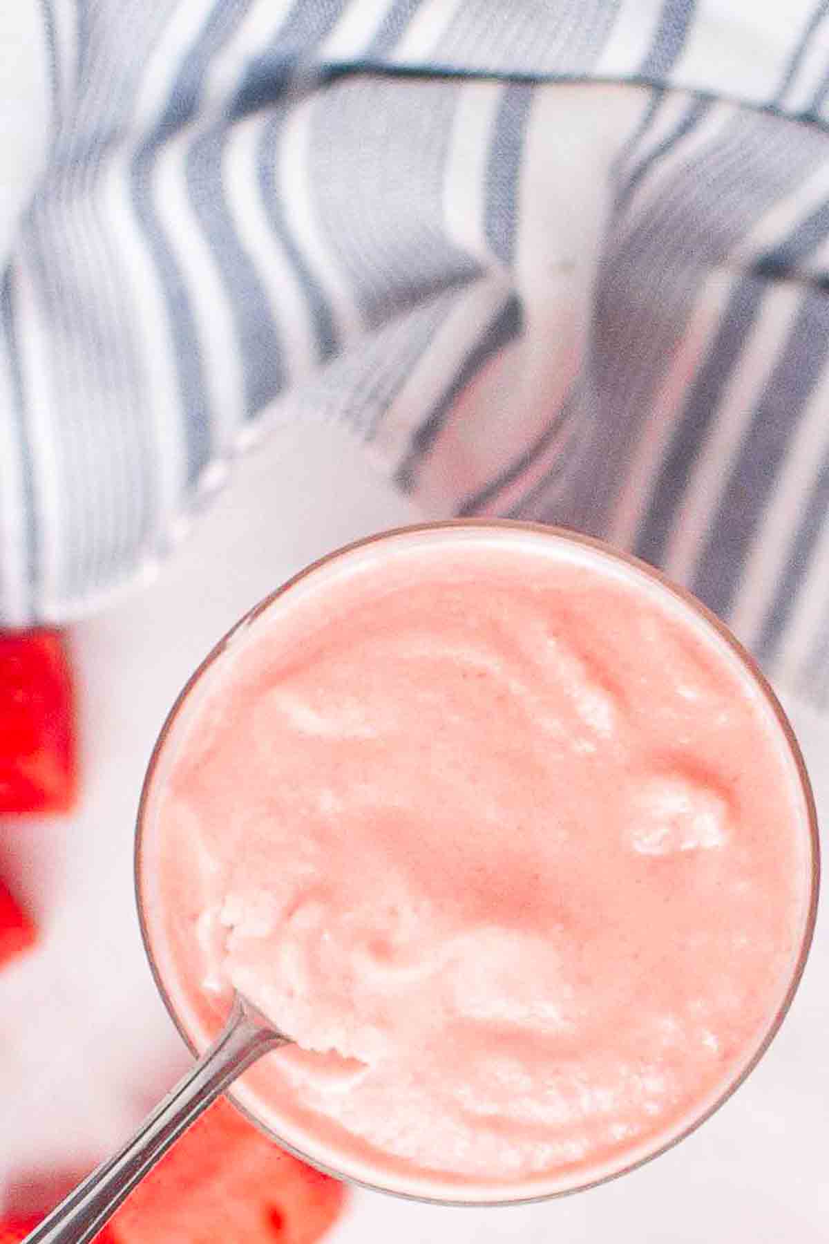 A glass of pink smoothie with a spoon in it, placed on a white surface next to a blue-and-white striped cloth.
