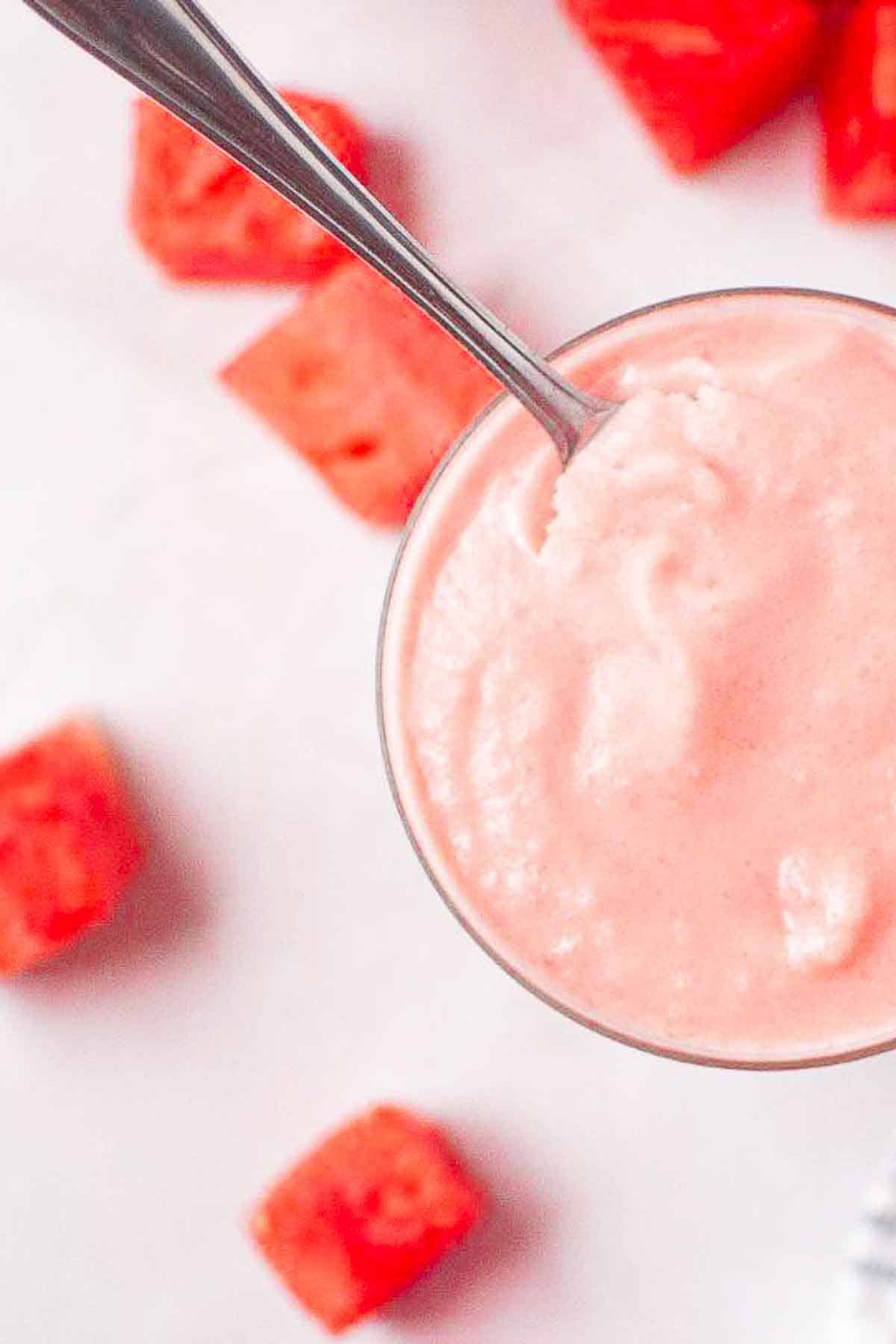 A spoon in a glass of pink smoothie, with watermelon cubes scattered on a white surface nearby.