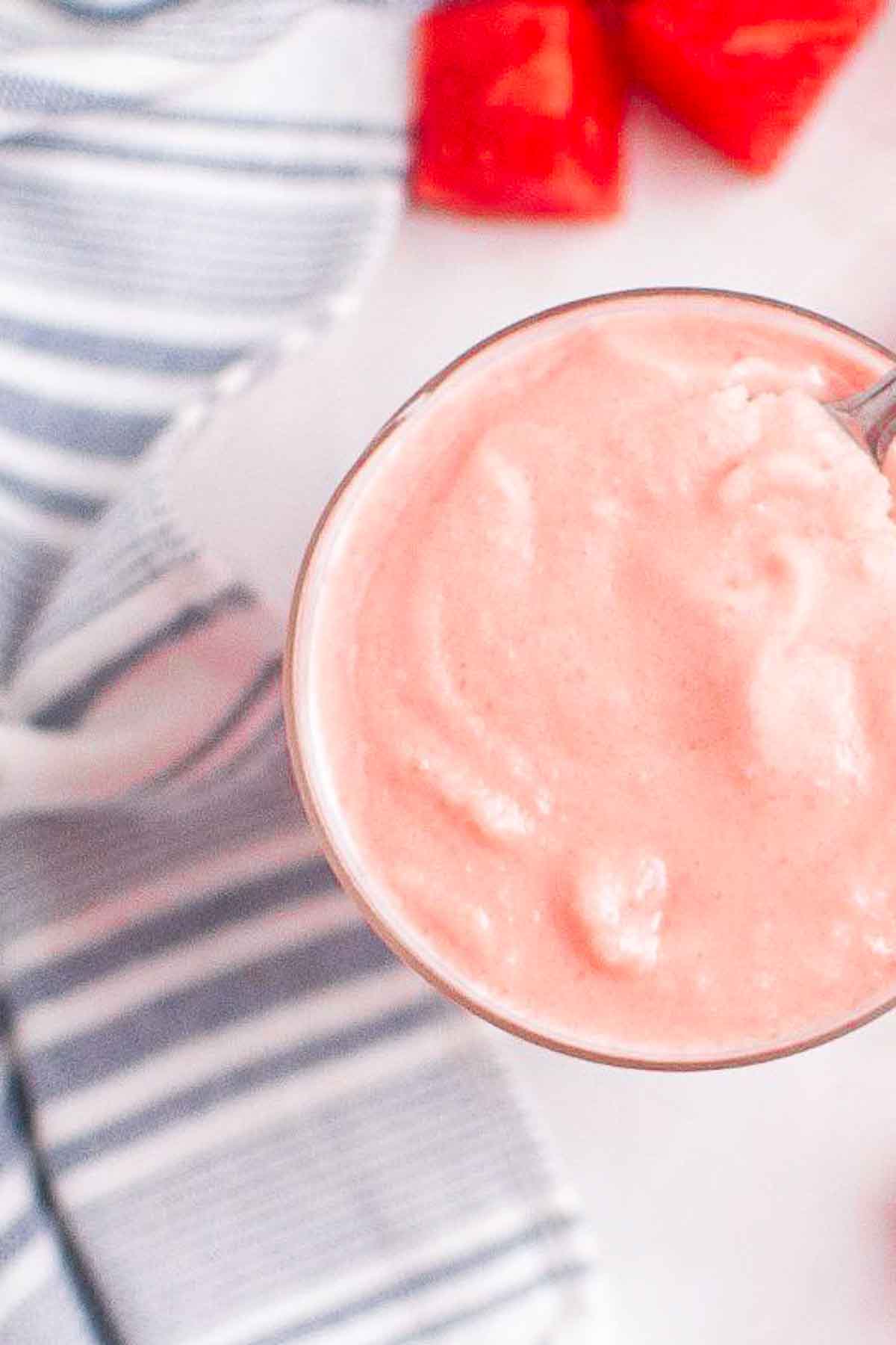 A glass filled with a thick pink smoothie sits on a white surface next to a striped cloth and a few pieces of cut watermelon.