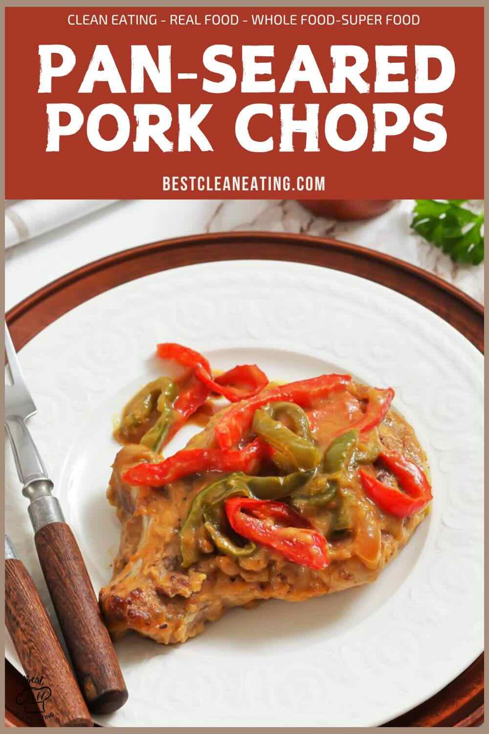 A pan-seared pork chop topped with sliced red and green peppers on a white plate with a brown border, next to a fork and knife.