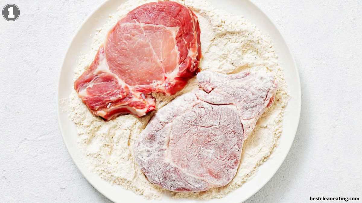 Two raw pork chops on a plate with flour; one chop is fully coated in flour, while the other is only partially coated.