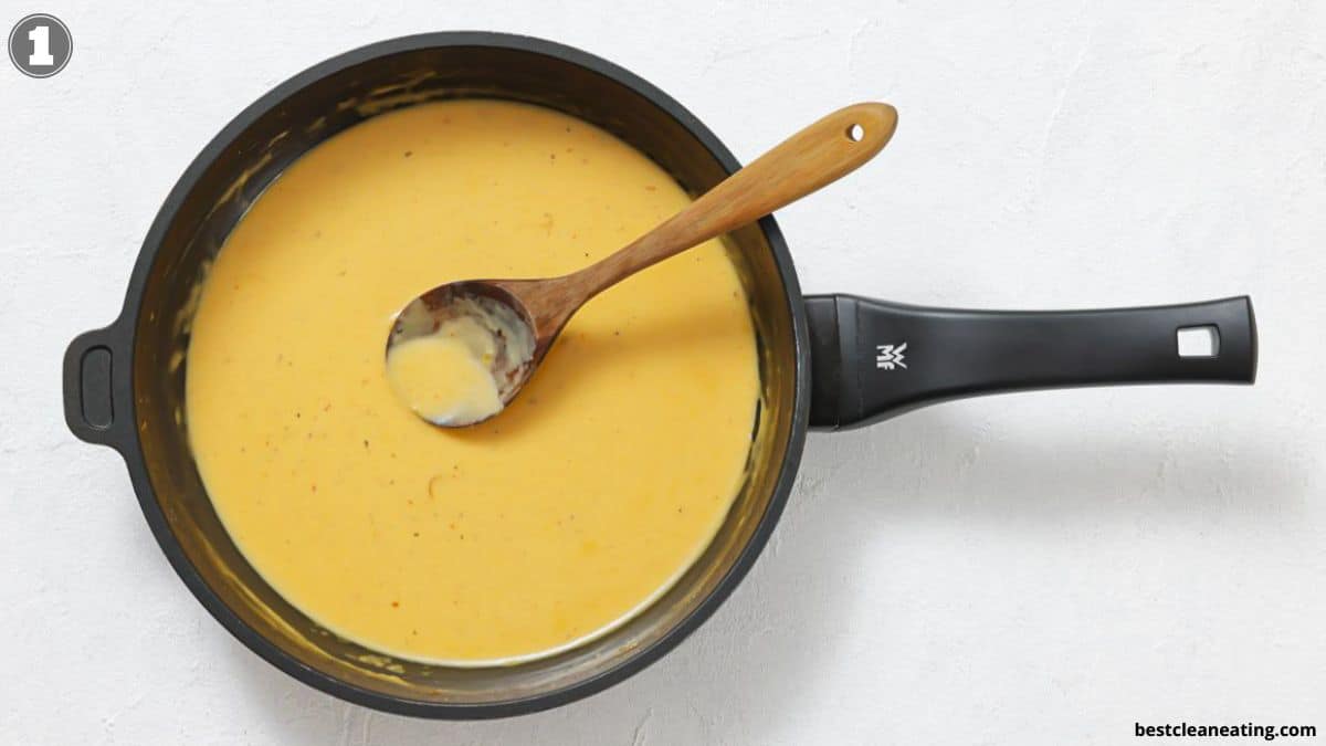 A saucepan filled with creamy yellow cheese sauce and a wooden spoon resting inside, placed on a white surface.