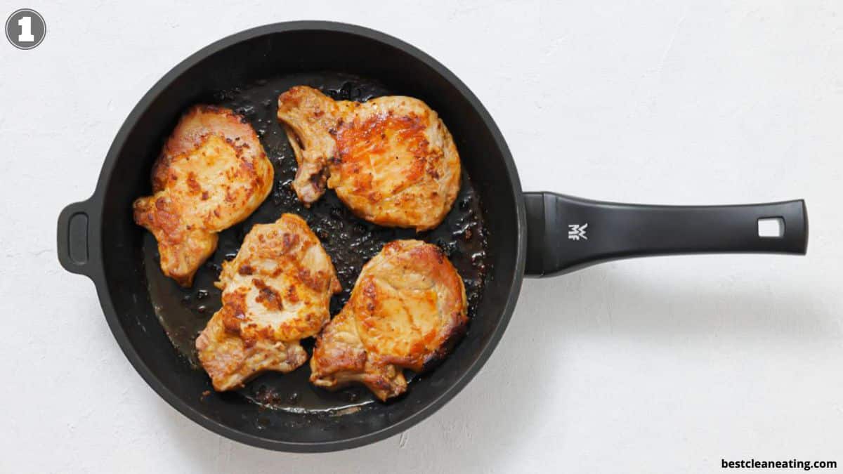 Four browned pork chops cooking in a black frying pan on a white surface.