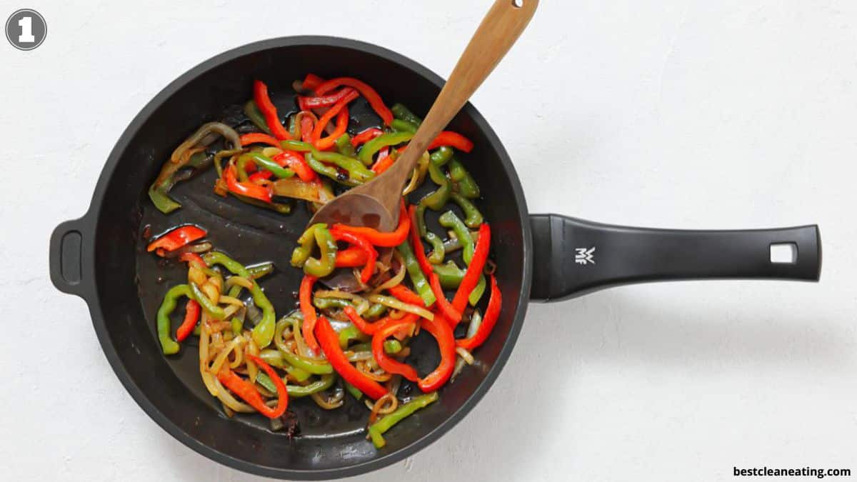 Sliced red and green bell peppers and onions being sautéed in a black frying pan with a wooden spoon.