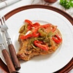 A cooked pork chop topped with sliced red and green bell peppers and sauce, served on a white plate with a fork and knife.