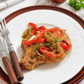 A cooked pork chop topped with sliced red and green bell peppers and sauce, served on a white plate with a fork and knife.